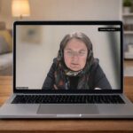 ufgeklapptes Notebook auf einem Holztisch im Wohnzimmer. Auf dem Bildschirm ist eine Videokonferenz zu sehen, in der eine Frau mit Headset und Brille spricht; eingeblendeter Name: „Andrea Lübcke“. Im Hintergrund sind Sofa, Lampe und Regal unscharf zu erkennen.